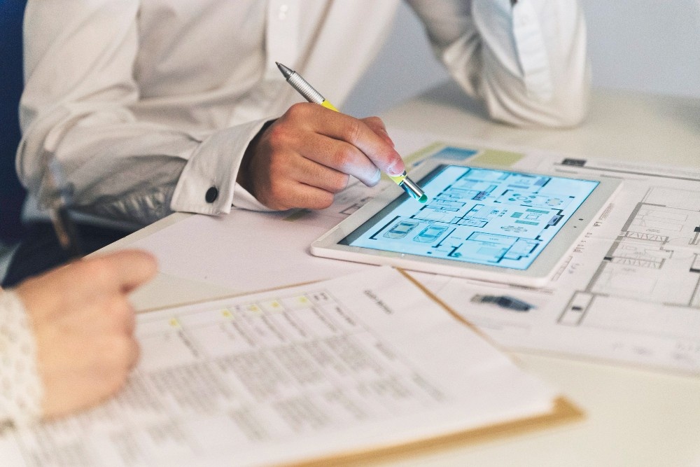 Two people review architectural blueprints on a tablet and paper. One of the two BIM Architects, points at the tablet with a pen. 
