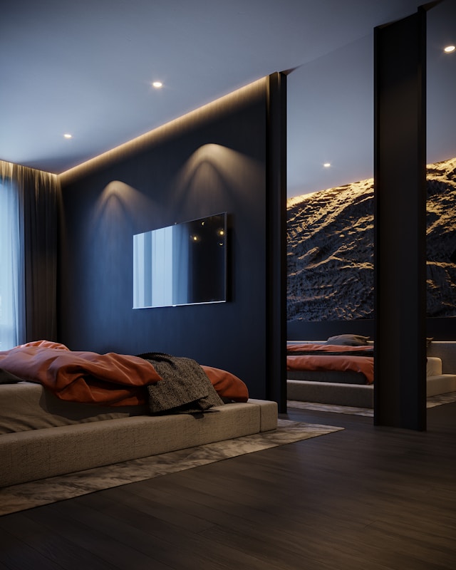 A modern bedroom showcasing innovative design solutions with a dark color scheme. The room features a bed with orange bedding, a large wall-mounted TV, and ambient lighting.
