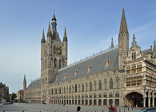 A large, historic Gothic building with a tall clock tower and ornate stonework stands under a clear blue sky. 