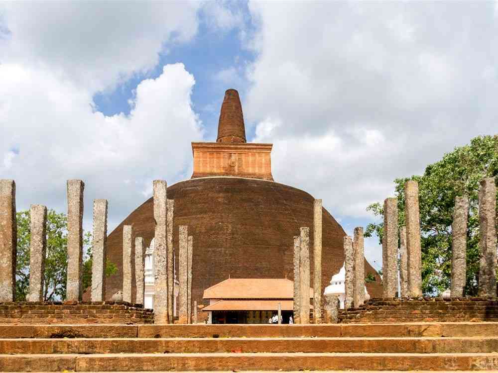 An ancient stupa, characteristic of Sri Lankan temple architecture, boasts a large domed structure and a towering central spire. Encircled by stone columns and steps, the scene reveals cultural insights with partly cloudy skies and trees visible on the right side.