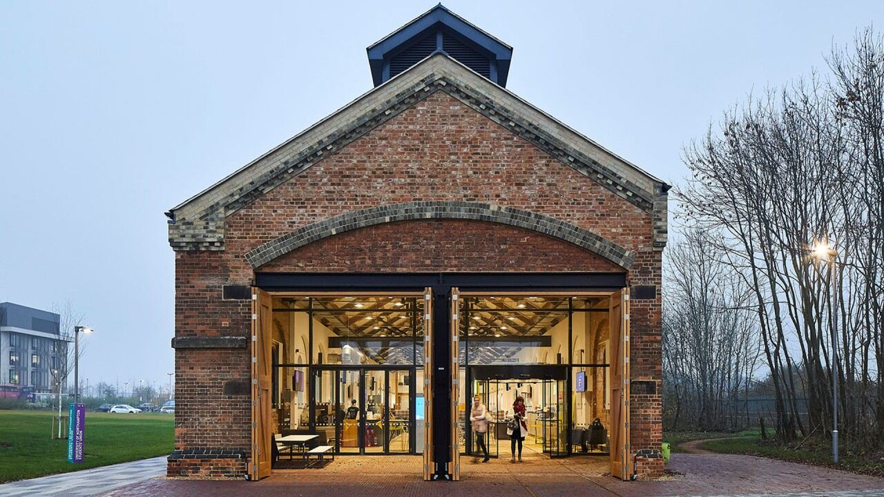 A brick building with a peaked roof and large open doors reveals a well-lit interior bustling with people. This fine example of adaptive reuse is surrounded by grass and trees, set against overcast skies, illustrating a commitment to sustainability in urban planning.