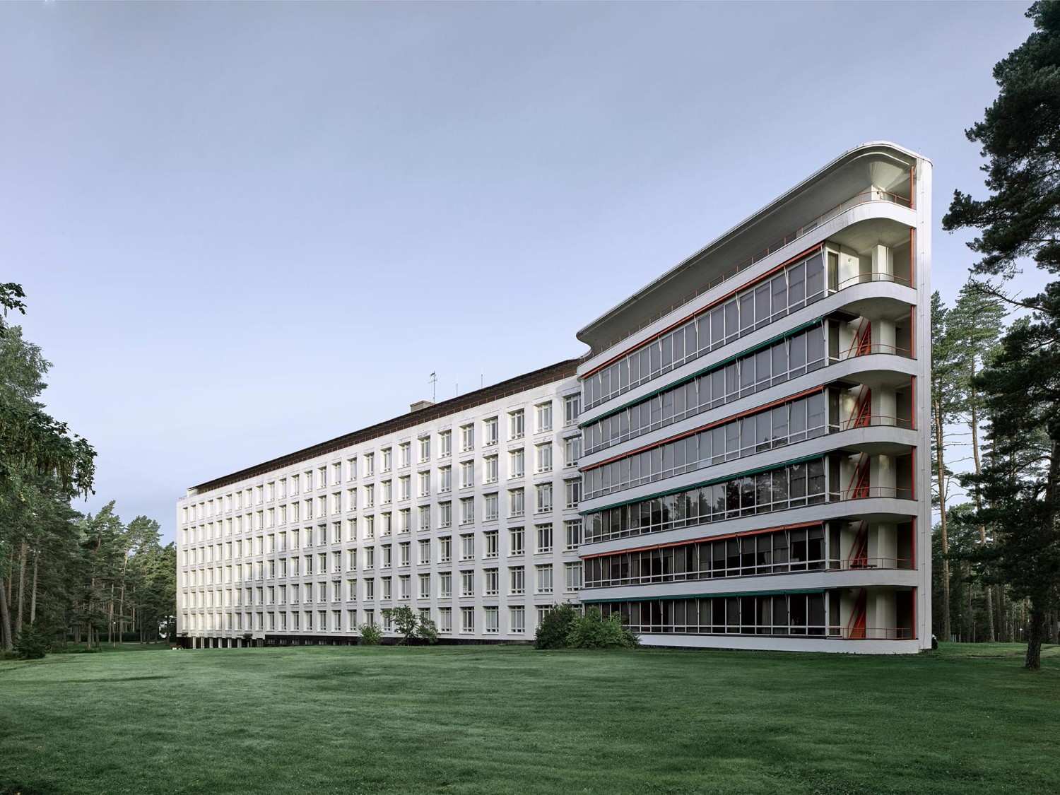 The modernist, Paimio Sanatorium building designed by visionary architect, Alvar Aalto, stands amidst a green lawn and surrounding trees. The structure features vertical rows of windows and a curved corner with balconies, embodying excellence in the built environment against a clear sky.