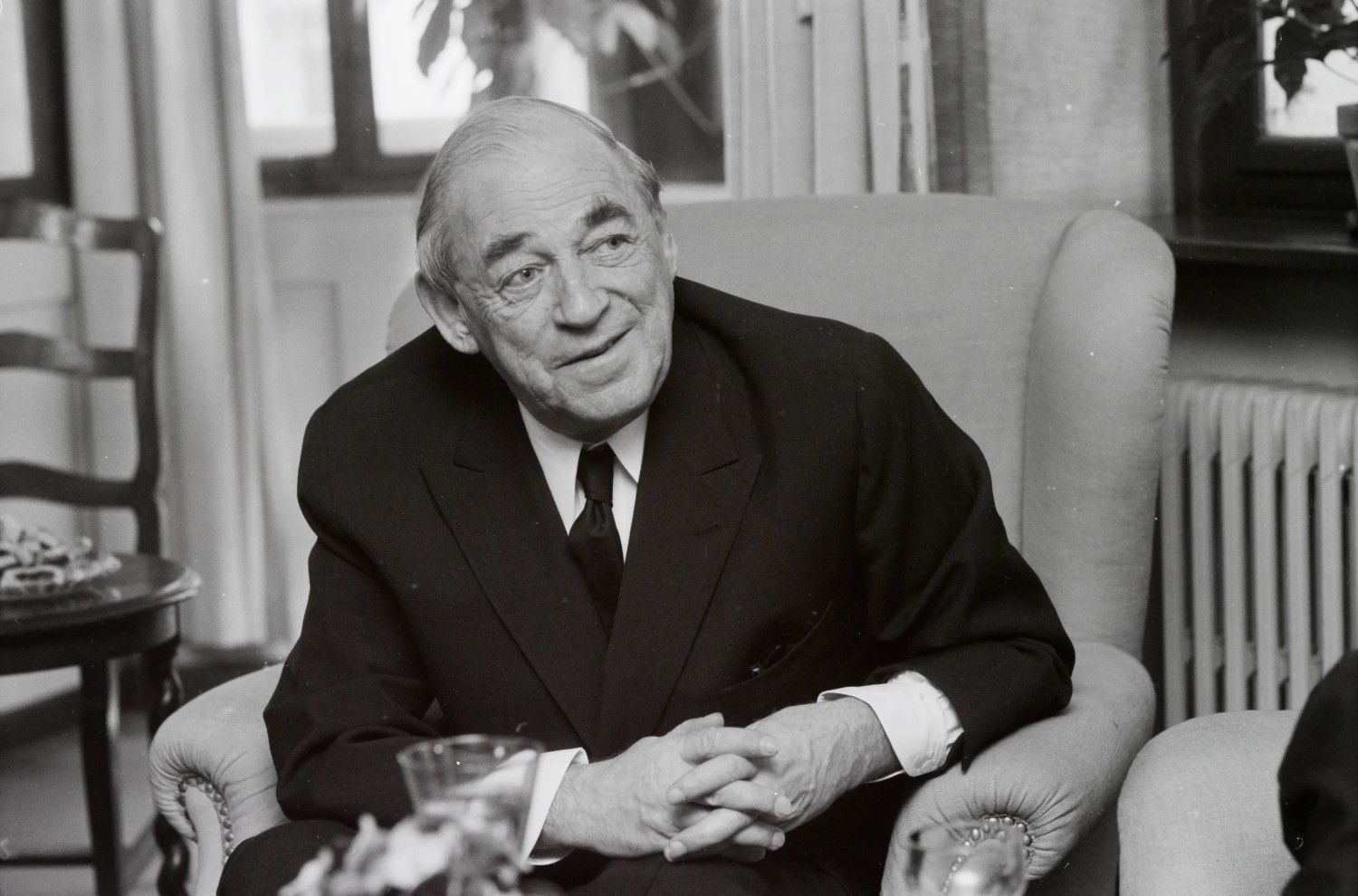 Alvar Aalto in a suit, sits in an armchair, smiling slightly with hands clasped. 