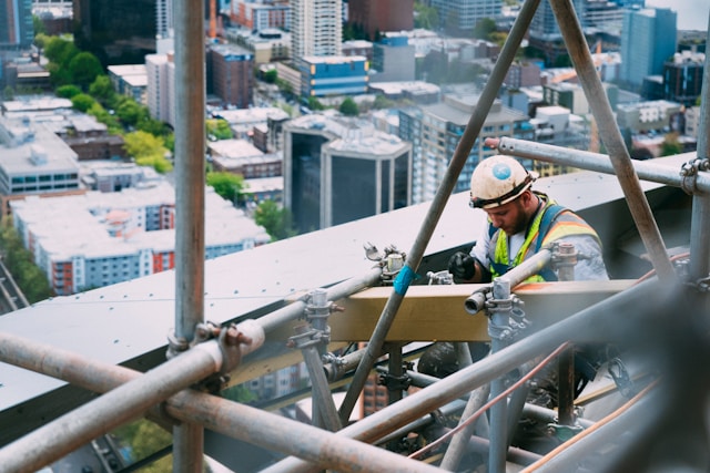 A construction worker wearing a hard hat and high-visibility vest demonstrates exceptional project management skills while working on scaffolding high above a cityscape. 