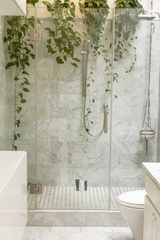 The glass shower enclosure featuring a rainfall showerhead is complemented by greenery hanging above, introducing a natural touch. With walls showcasing a light marble texture and thoughtful toilet design, there's also a white bathtub nearby. 