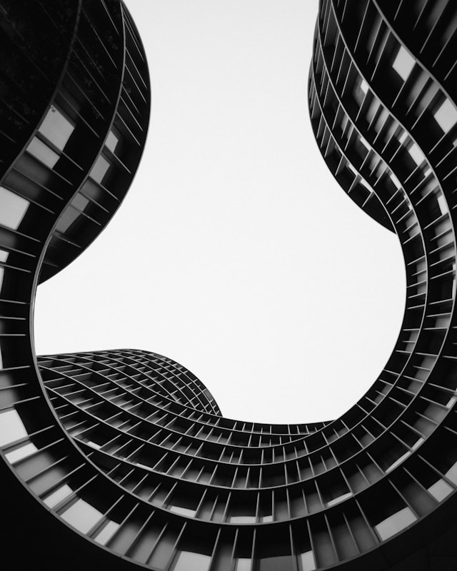 Black and white image of a modern, wavy architectural structure viewed from below. The curving lines form an abstract pattern against the sky, showcasing the building's unique design and geometric windows, a testament to sustainable architecture.