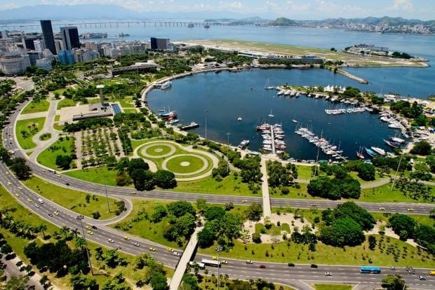 Aerial view of a large city park with green spaces and winding roads surrounding a serene bay filled with docked boats. Tall buildings and a long bridge, an engineering marvel, can be seen in the background, with mountains and water stretching out to the horizon.