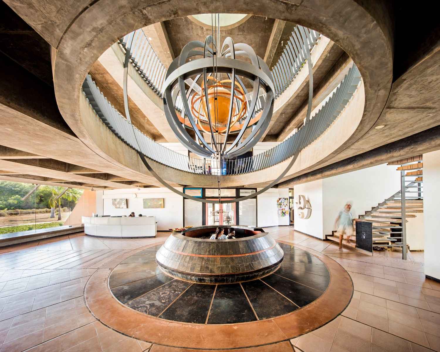A spacious interior features a central, intricate metallic sculpture suspended from the ceiling above a circular fountain. It is a sustainable building in Auroville, India.