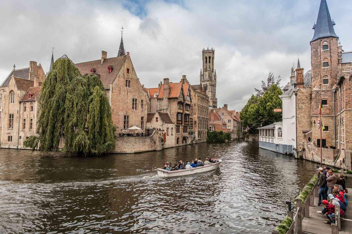 A scenic view of a canal in Bruges, Belgium, showcases Gothic urbanism as a boat carries passengers past historic brick buildings and a tall church tower. 
