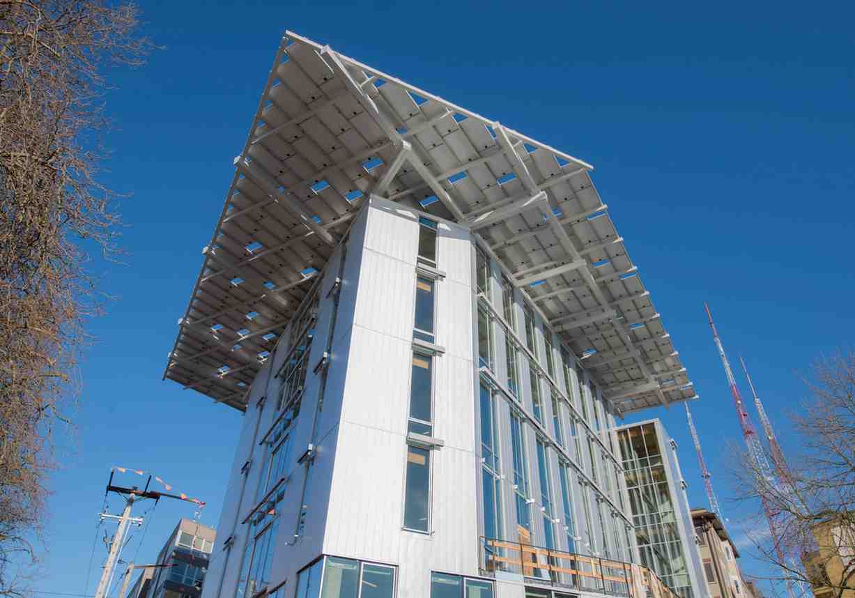 A modern green building features an angular design with extensive solar panels on the roof against a clear blue sky. The structure, showcasing sustainable strategies, has large glass windows and is surrounded by trees and other structures.