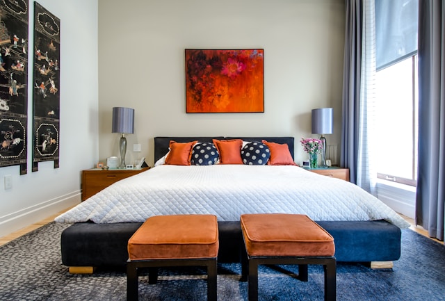 A modern bedroom adhering to Vastu architecture features a king-sized bed with a white quilt, orange and polka dot cushions. 