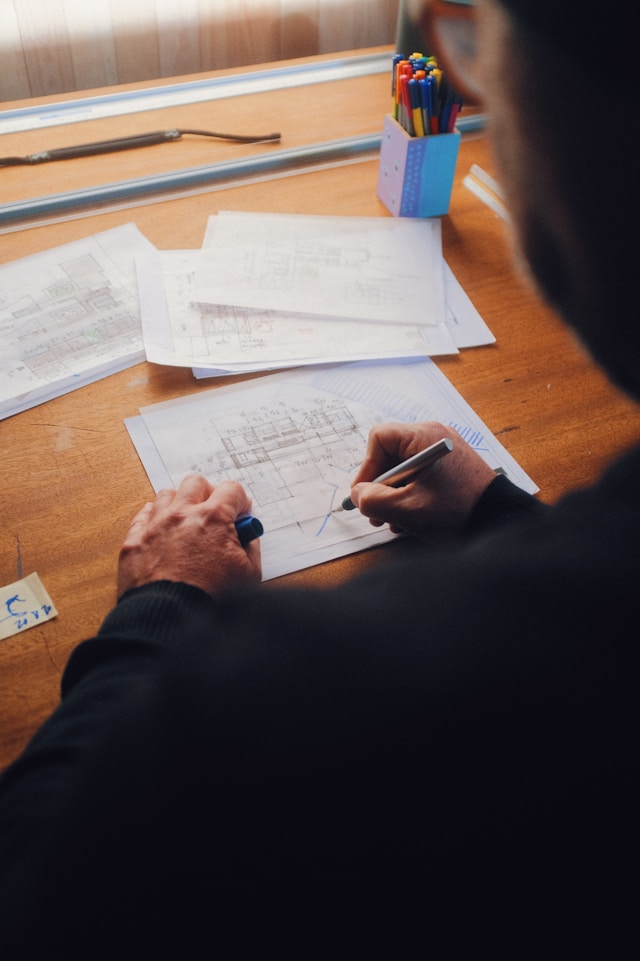 A person is writing on a sheet of paper with architectural drawings spread across a wooden desk, hinting at collaborative design.&nbsp;