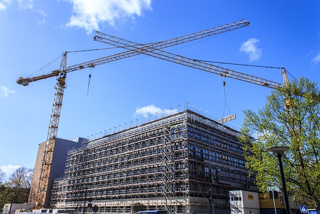 A construction site with two large cranes crossing above a multi-story building draped in scaffolding showcases the architects' collaboration. 