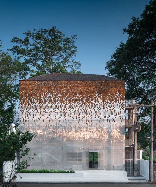 A modern building with a unique facade design transitions from a wooden shingle texture on top to a reflective, shimmery surface at the bottom, surrounded by trees under a clear blue sky. 