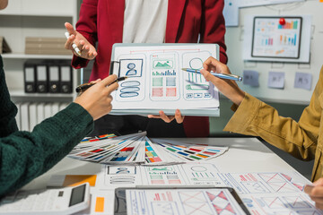 Three people are engaged in a Design Thinking session at a table, discussing website design mockups. One person holds up a visual layout, while others point at details with pens.