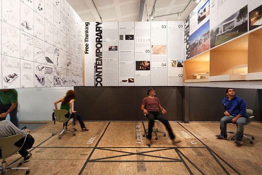 A group of people sitting on swivel chairs in a parametric design exhibition space featuring architectural designs and photographs on the walls. The floor is marked with lines and words.