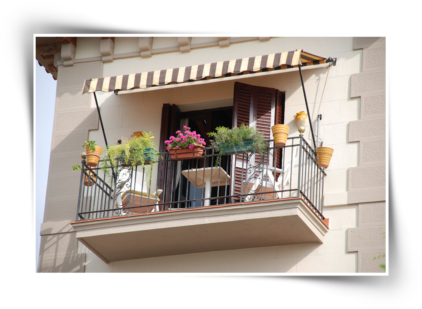Small residential balcony with striped awning, wrought iron railing, potted plants, outdoor seating, and wooden shutters