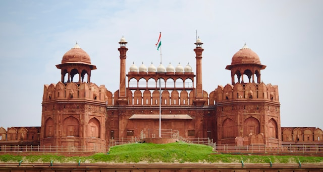 A large, historic fort crafted from red sandstone showcases striking Mughal architecture with its domes and arched structures. An Indian flag flutters proudly at the front against the clear blue sky.