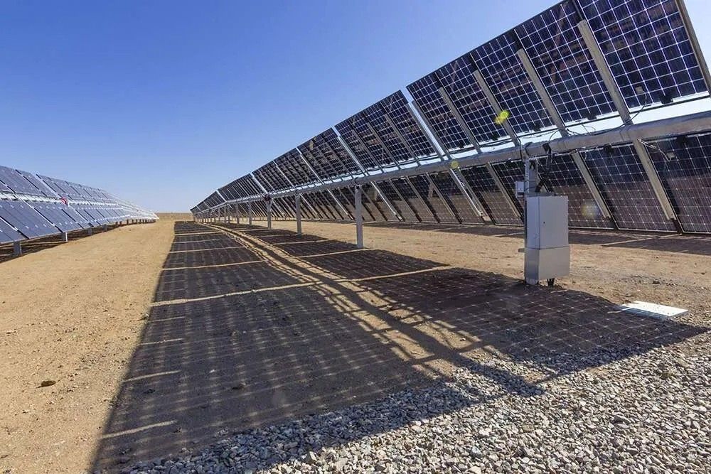 Rows of solar panels, an eco-friendly structure nestled in the desert landscape, are angled towards the sky with clear blue overhead. Shadows cast on the sandy ground illustrate a large-scale solar energy facility dedicated to renewables.