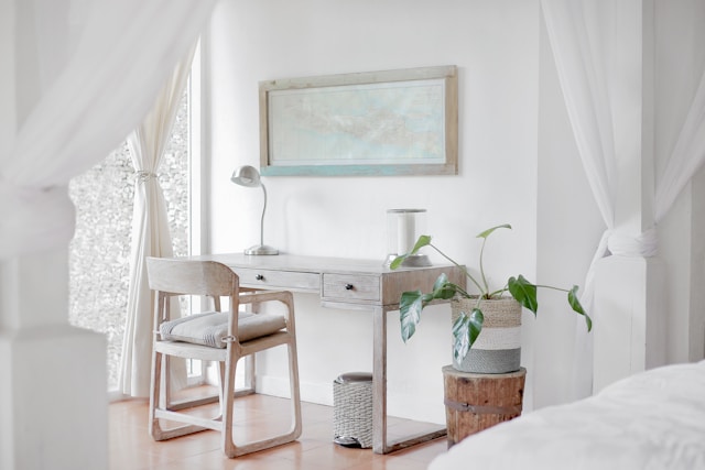 A bright, minimalist bedroom showcases a white desk paired with a wooden chair. Influenced by Vastu principles, a lamp and potted plant rest on the desk beneath a framed map.&nbsp;