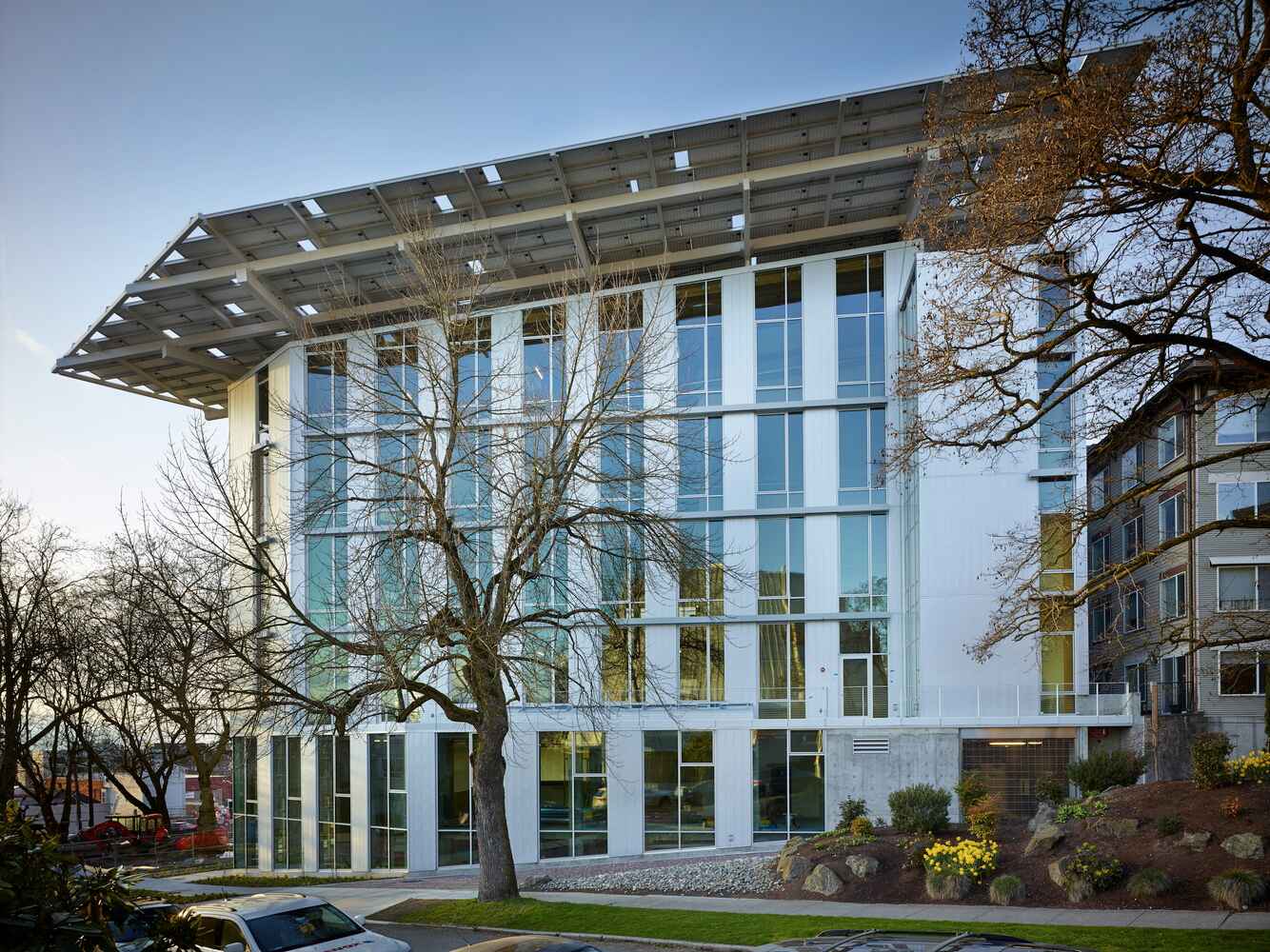 A modern, multi-story building with large glass windows and a prominent overhanging roof sits among trees and landscaping. Embracing sustainable architecture, its design maximizes natural light while casting shadows. 