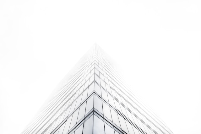 A tall, modern skyscraper with a glass facade disappears into a white, foggy sky, creating a minimalistic and ethereal effect. Emphasizing its towering presence, this masterpiece of sustainable architecture features sleek lines and reflective surfaces that adapt to the environment.