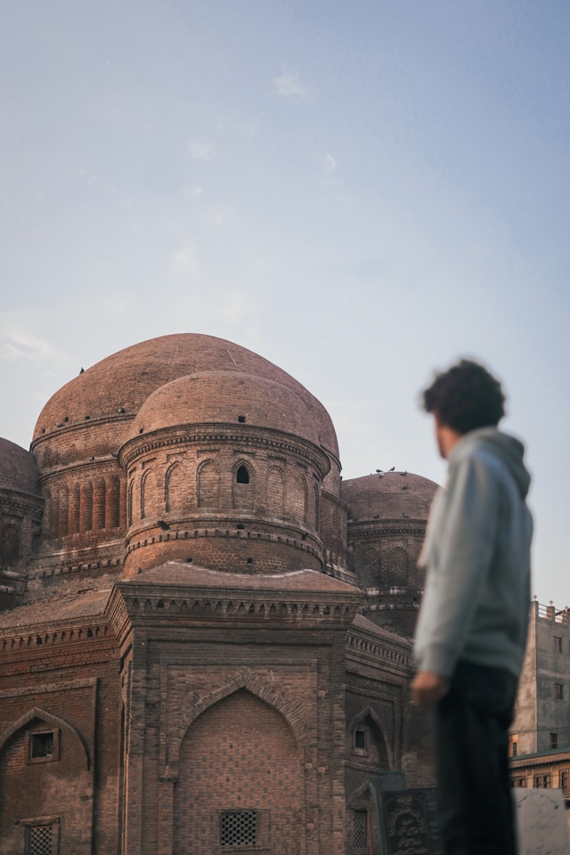 A person stands in the foreground, gazing at a historical brick building with large domes against a clear sky. The architecture, reminiscent of Kashmiri styles, features intricate patterns and arches that echo the cultural impact of ancient Sufi shrines.