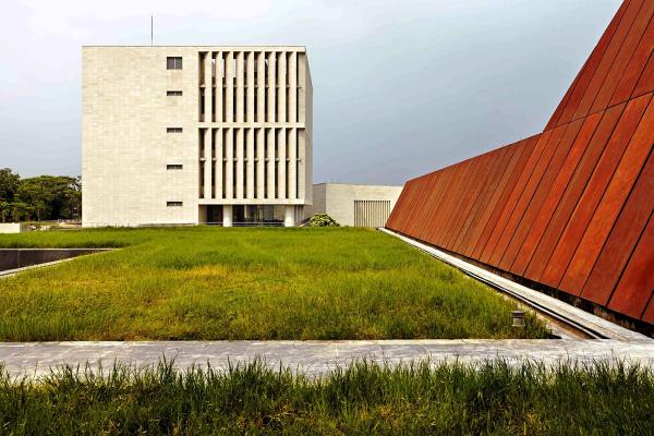 A modern building with vertical slits on its facade stands near a sloped, rust-colored structure, reflecting global styles.&nbsp;