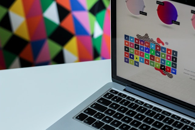 A laptop displaying colorful icons on its screen is positioned on a white surface. In the background, a vibrant geometric pattern with triangles enhances the lively and artistic ambiance, reflecting the essence of collaborative design in innovative spaces.