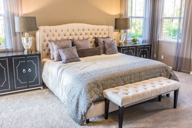 A cozy bedroom featuring a large, tufted bed with neutral bedding exudes positive energy. Natural light streams through two windows, creating a warm and inviting atmosphere steeped in vastu design principles.