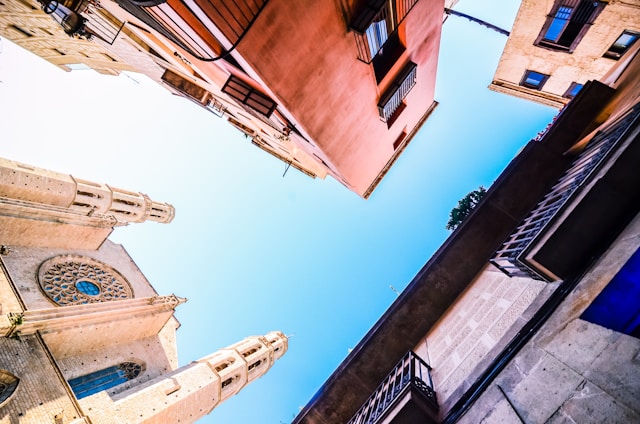 View of tall historical buildings and a church from a low angle against a clear blue sky. The universal architecture showcases detailed stonework with contrasting warm and cool tones. 