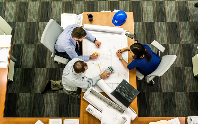 From an aerial view, three people at a desk delve into architectural plans, one with a calculator, another with a pen, and the third noting down ideas. 