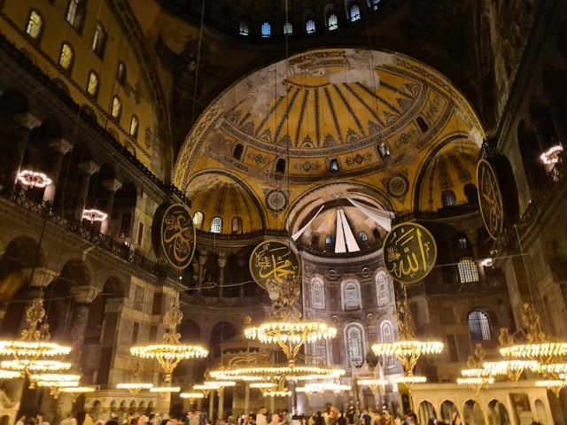 The interior of this grand, historic building boasts large, ornate domes elegantly supported by pendentives, with intricate decorations. 