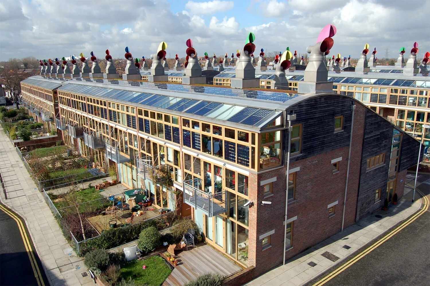 A row of modern, net zero energy townhouses features solar panels on the roofs and colorful wind turbines. Each unit has a small garden, while renewables like these line the greenery-filled street. 