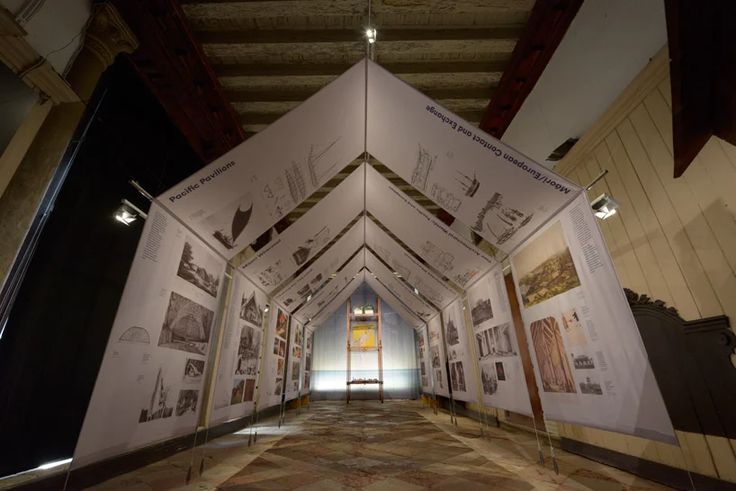 A modern art exhibition features a tent-like structure with printed panels displaying architectural drawings and photos. Utilizing parametric design, it stands on a tiled floor in a dimly lit room with exposed wooden beams on the ceiling.