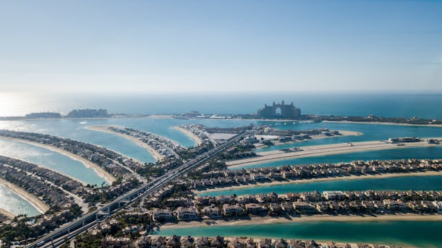 Aerial view of the Palm Jumeirah in Dubai, a landfill project, showcasing the marvel of urban planning with man-made islands resembling a palm tree. The islands are lined with residences and surrounded by turquoise water. 
