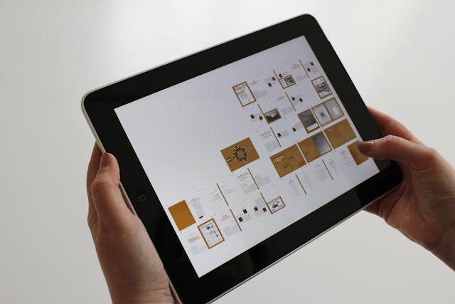A person holds a tablet displaying various UX design mockups with a minimalist layout. The screen shows multiple small images and text blocks on a white background, creating an immersive experience. The backdrop is plain and softly lit.