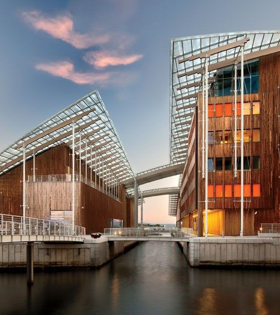 Modern buildings with wooden facades and glass roofs stand on either side of a canal.&nbsp;