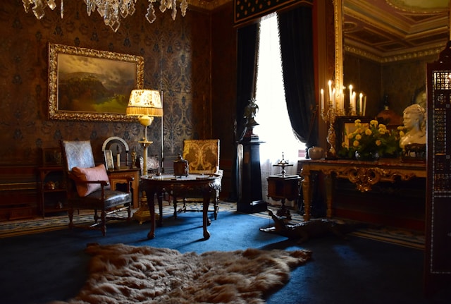 A lavish, dimly lit room with ornate wallpaper, antique wooden furniture, and a chandelier. 