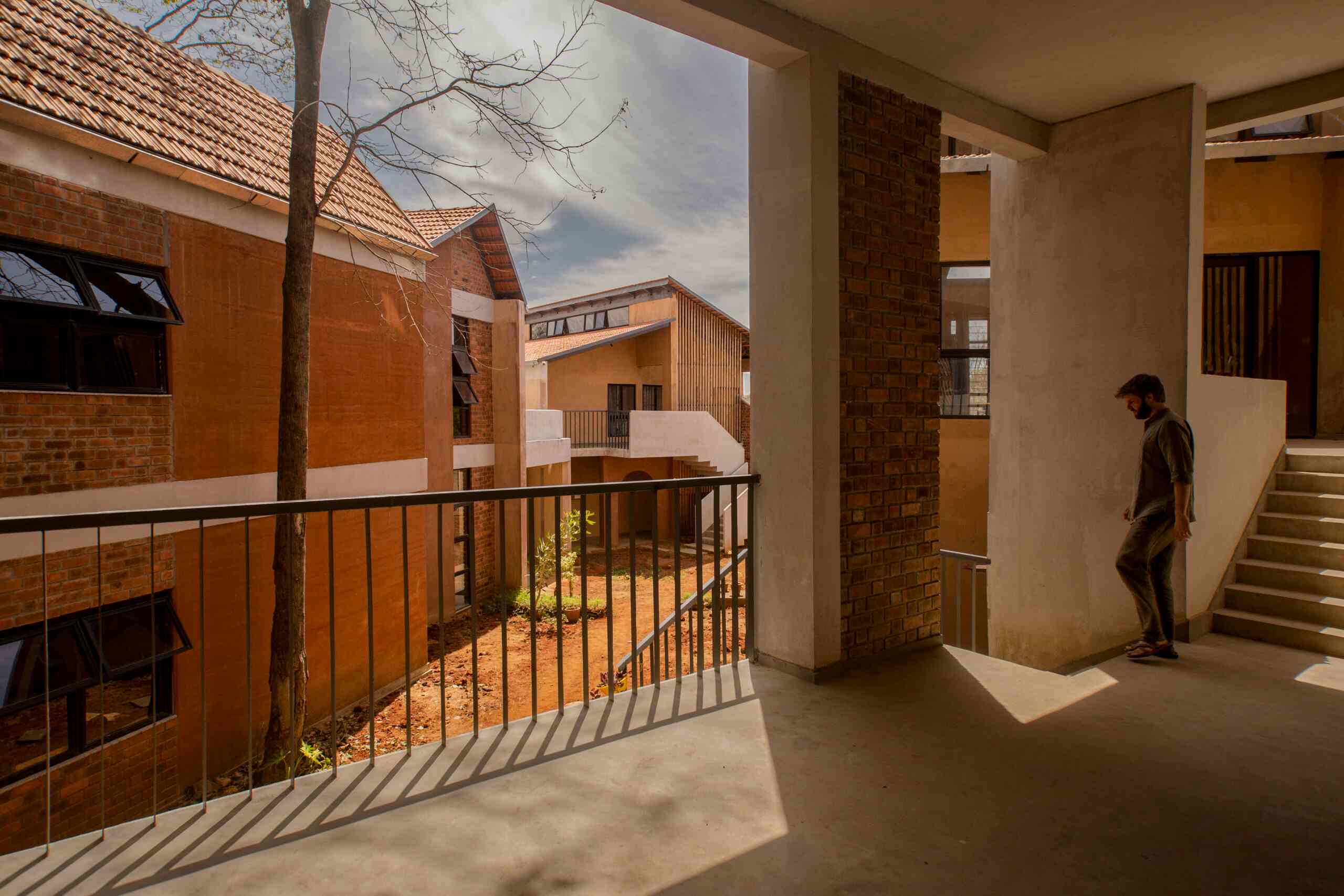 A man stands on a sunlit balcony overlooking a courtyard, designed with sustainability in mind. The area is encircled by modern brick and concrete buildings with slanted roofs and various windows, showcasing the architects' vision. 