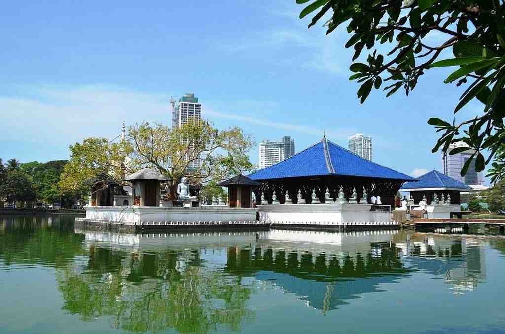 A serene lake with reflective water features a temple of Sri Lankan architecture, with blue roofs and white walls, surrounded by trees.&nbsp;