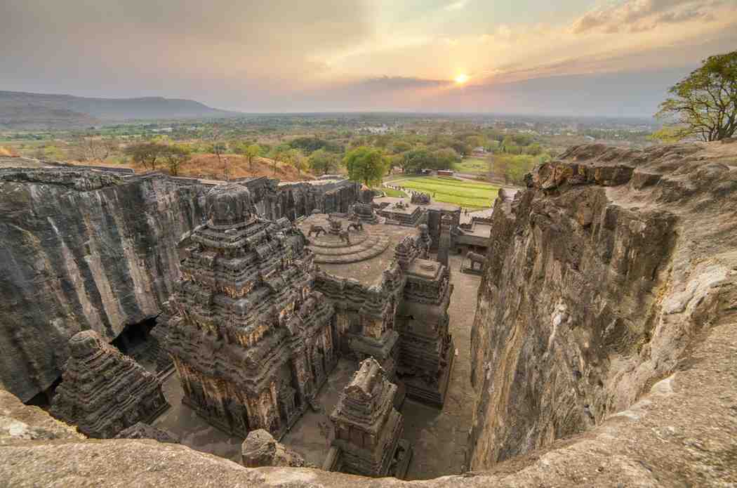 An aerial view of the ancient Kailasa Temple, carved into rock, at Ellora Caves in India. This exquisite example of Maharashtrian architecture is encircled by rocky cliffs, offering a vast landscape with a stunning sunset in the background.