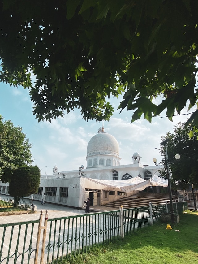 A serene white temple, echoing Kashmiri architecture with its large dome, sits under a bright blue sky, partially shaded by leafy green trees in the foreground. 