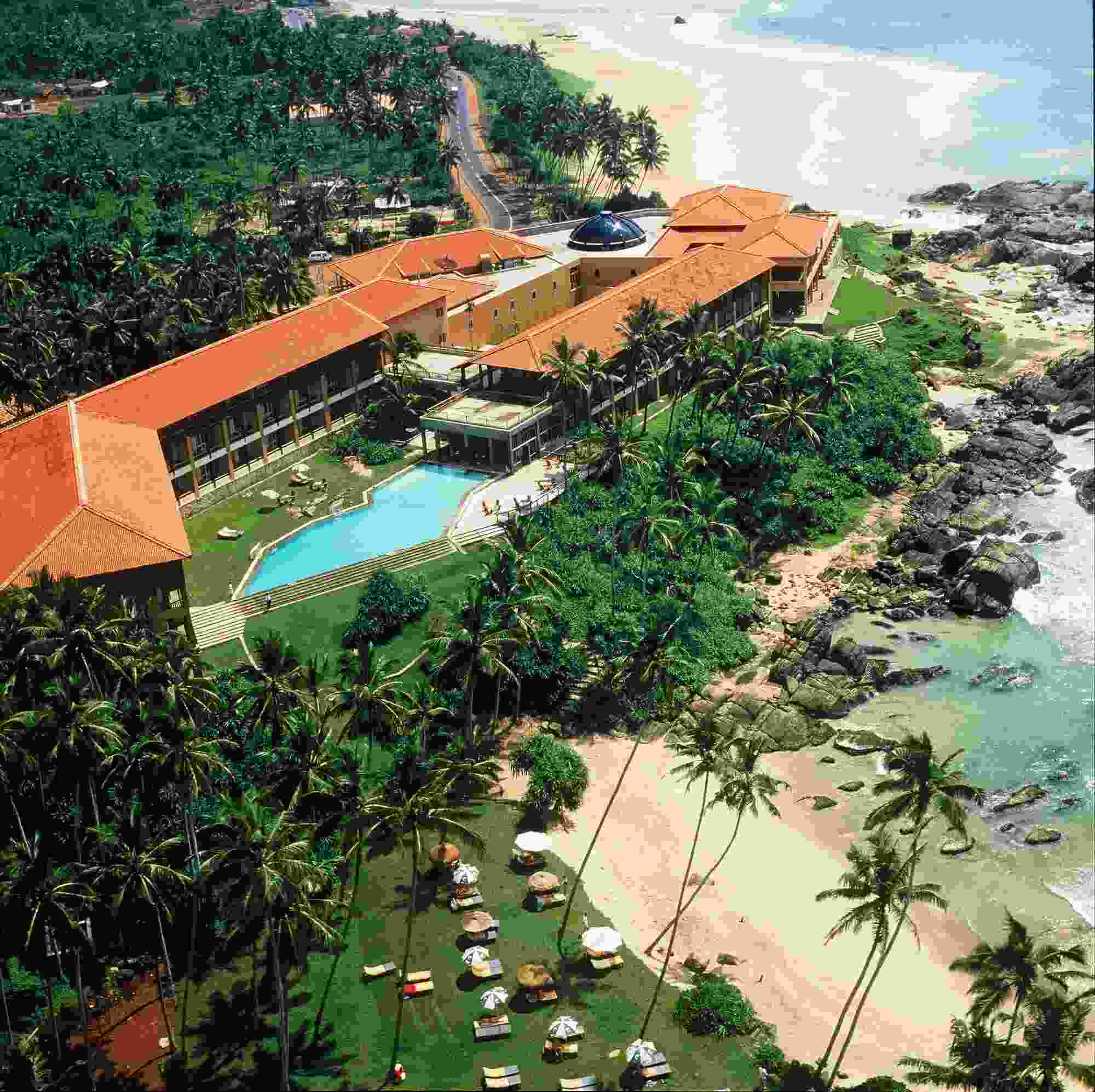 Aerial view of a coastal resort surrounded by palm trees. The resort, reflecting Sri Lankan architecture, features a large building with a red-tiled roof, a spacious swimming pool, sun loungers on a grassy area, and direct beach access with gentle waves and rocky outcrops along the shoreline.