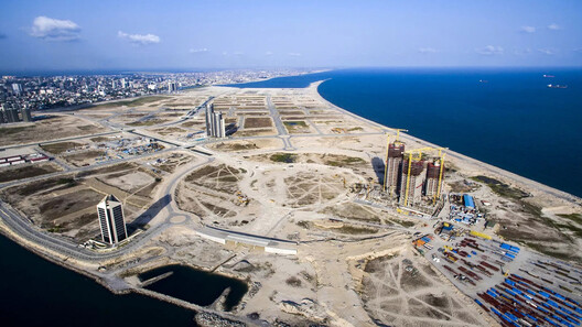 Aerial view of a coastal city with partially constructed buildings and vacant plots on a reclaimed land near the shoreline. The scene, an engineering marvel, captures a developing urban area adjacent to the ocean, with high-rise buildings and infrastructure under construction.