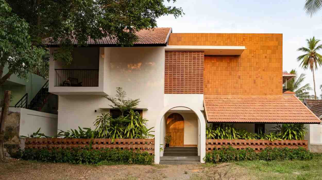 A contemporary house with a blend of white and orange exterior walls embodies Kerala architecture, featuring a tiled roof, an arched wooden door, and lush greenery in the front yard. 