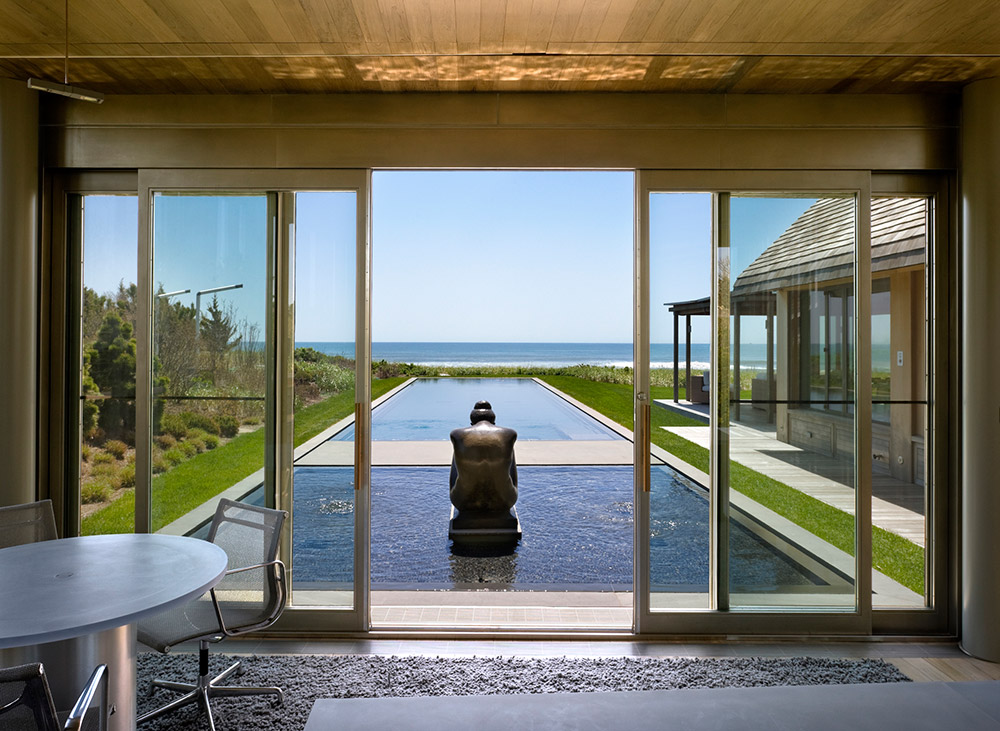 Through large glass doors, a modern room reveals a sculpture centered in a long reflective pool, flanked by greenery that highlights ecological planning. In the background, the ocean meets the horizon under a clear blue sky.