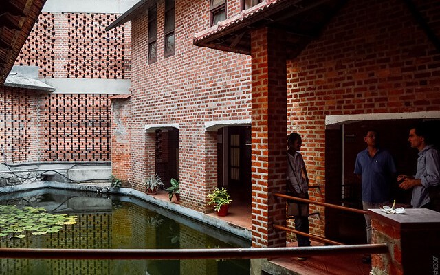 A group of three people stands in a courtyard with exposed brick walls. The space features a central pond with lotus plants, reflecting its sustainable design. The architectural style, characterized by open walls and a slanted roof, uses eco-friendly materials to maximize natural light.